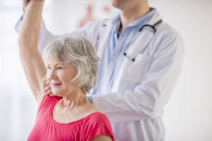 A senior women in a physical therapy session with her right arm raised overhead and assisted by a physical therapist.